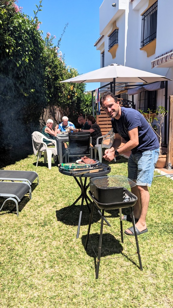 Een man staat bij een barbecue in een tuin, terwijl vrienden en familie aan een tafel zitten. De zon schijnt en er zijn ligstoelen in de achtergrond.
